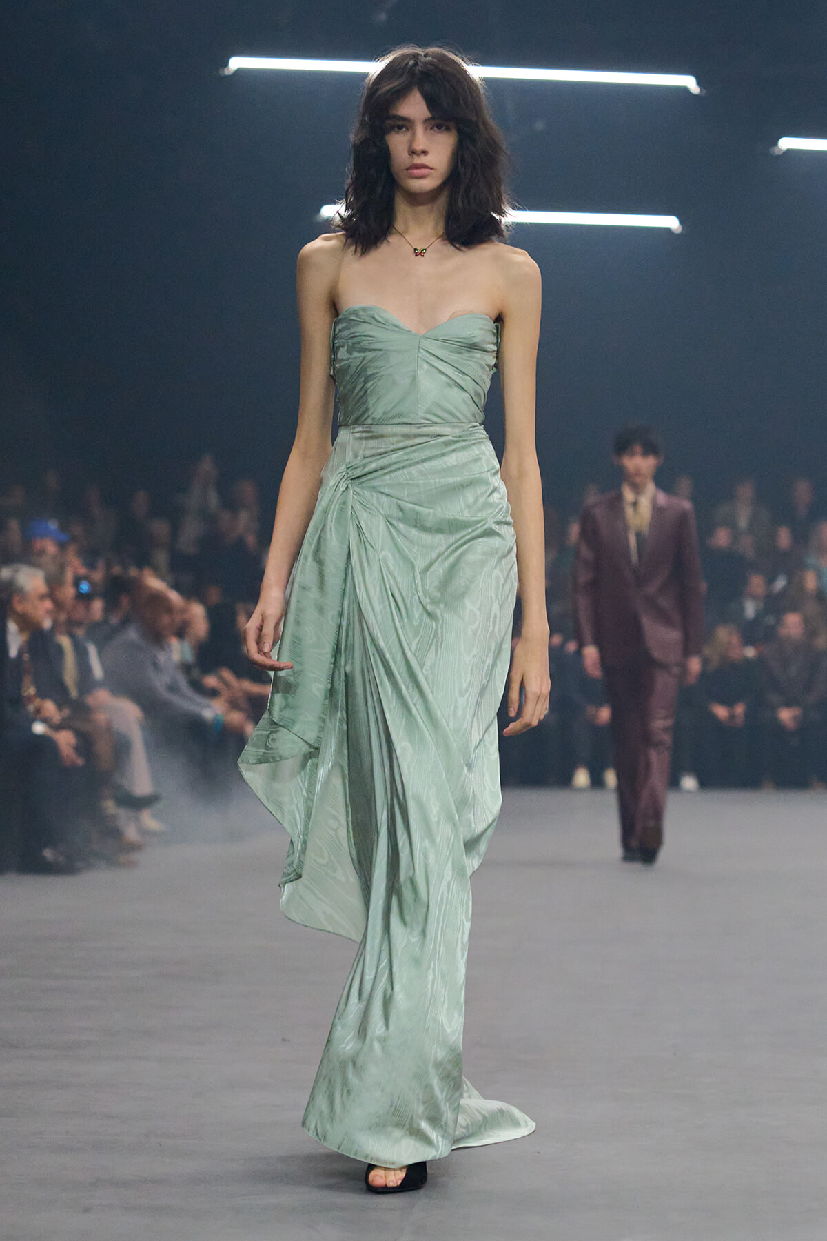 Model walks the runway in a strapless mint-green gown with a ruched bodice and flowing draped skirt, audience in the background.