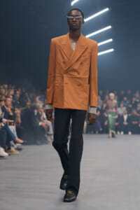 Male model walks the runway in an orange double-breasted blazer, black trousers, and glittery sunglasses with silver cuffs visible beneath the sleeves.