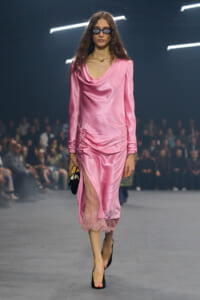 Model walking a fashion runway in a pink satin draped midi dress with a thigh-high slit and lace trim, paired with black heels and sunglasses.