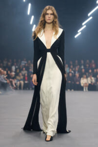 Runway model wearing a black-and-white gown with wide white lapels, a cinched waist, and flowing black panels; audience blurred in the background.