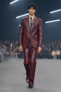 Male model walks a runway in a burgundy leather suit with a cream shirt and dotted tie; audience blurred in the background.