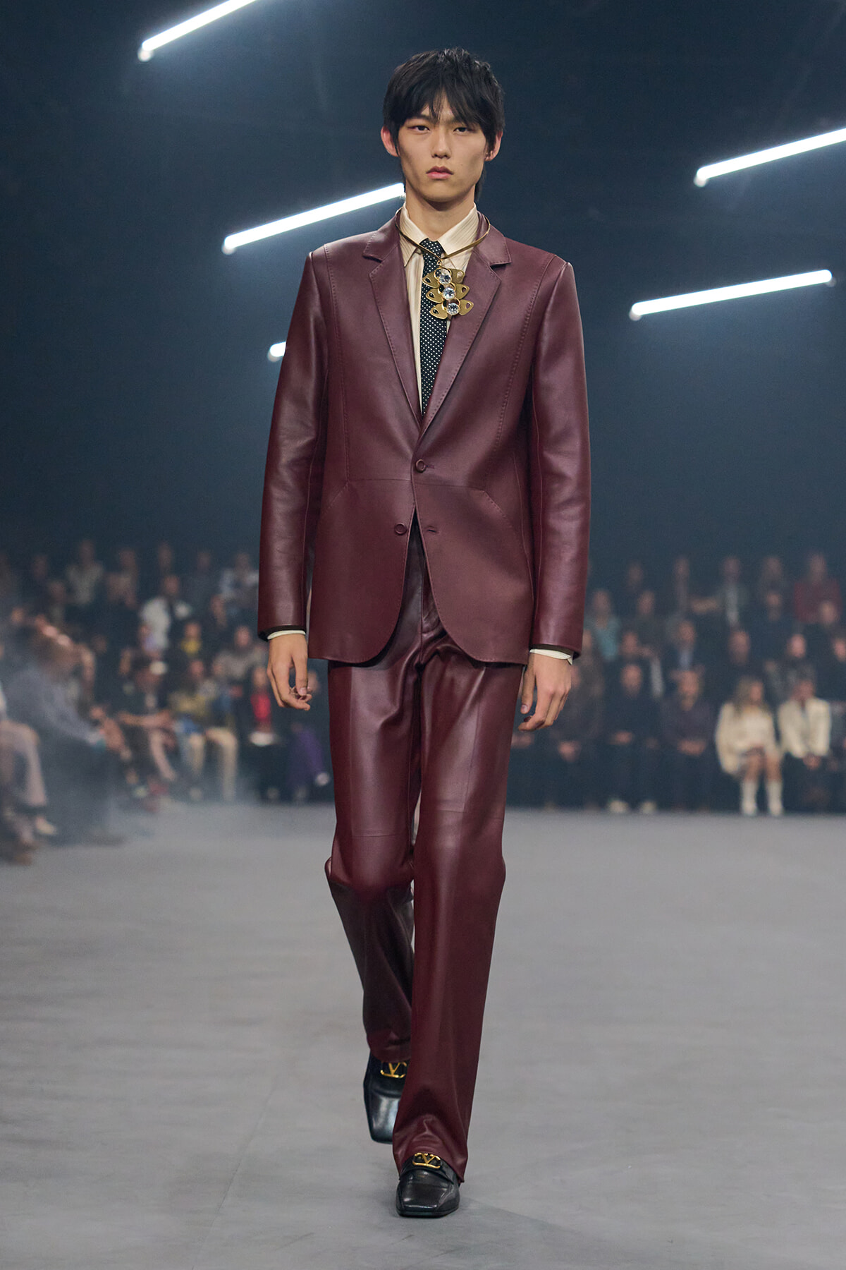 Male model walks a runway in a burgundy leather suit with a cream shirt and dotted tie; audience blurred in the background.