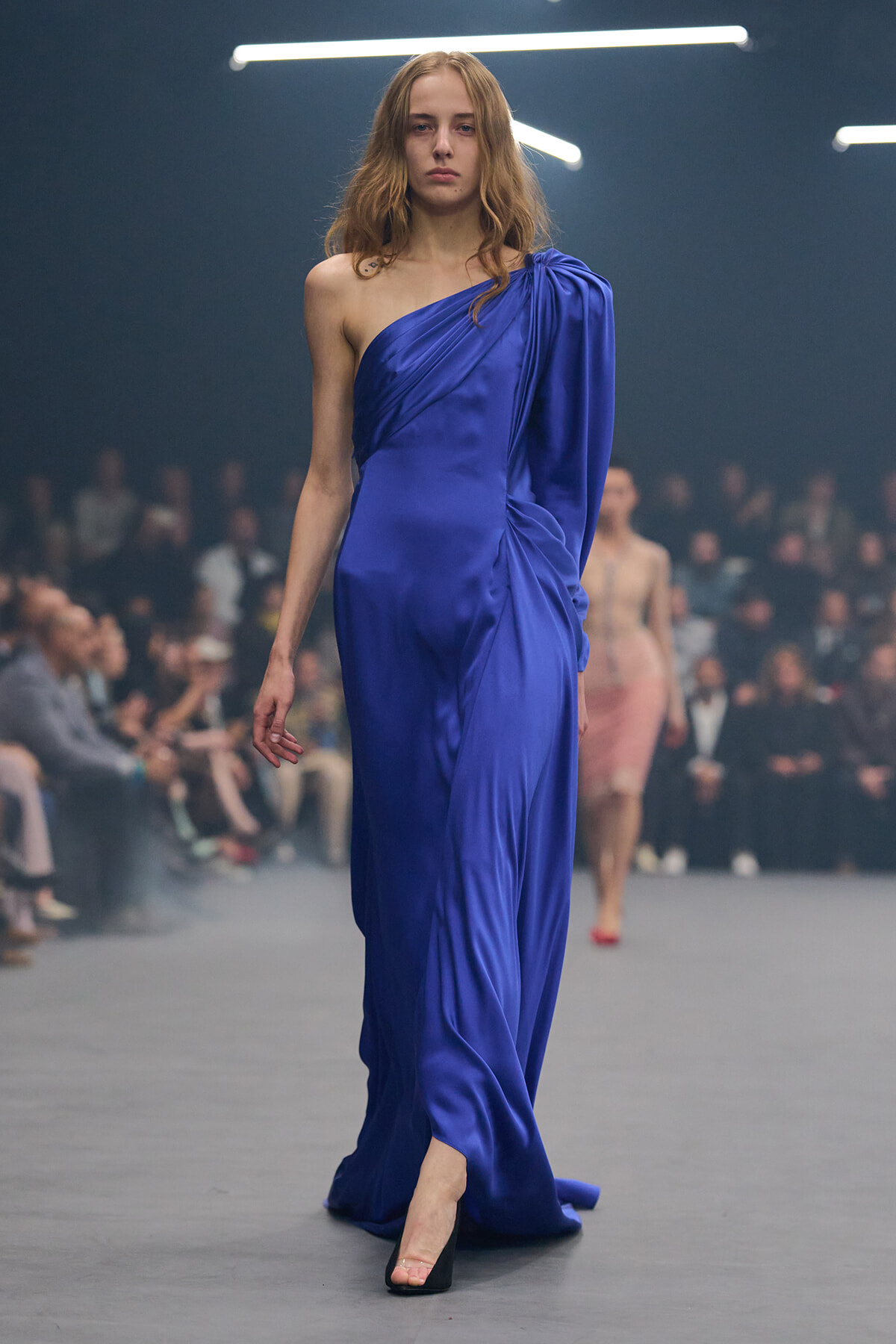 Model walking a runway in a one-shoulder royal blue satin gown, black heels, focused expression.