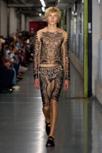 Male model walks the runway in a nude-toned sheer black lace bodysuit with ornate swirling embroidery.