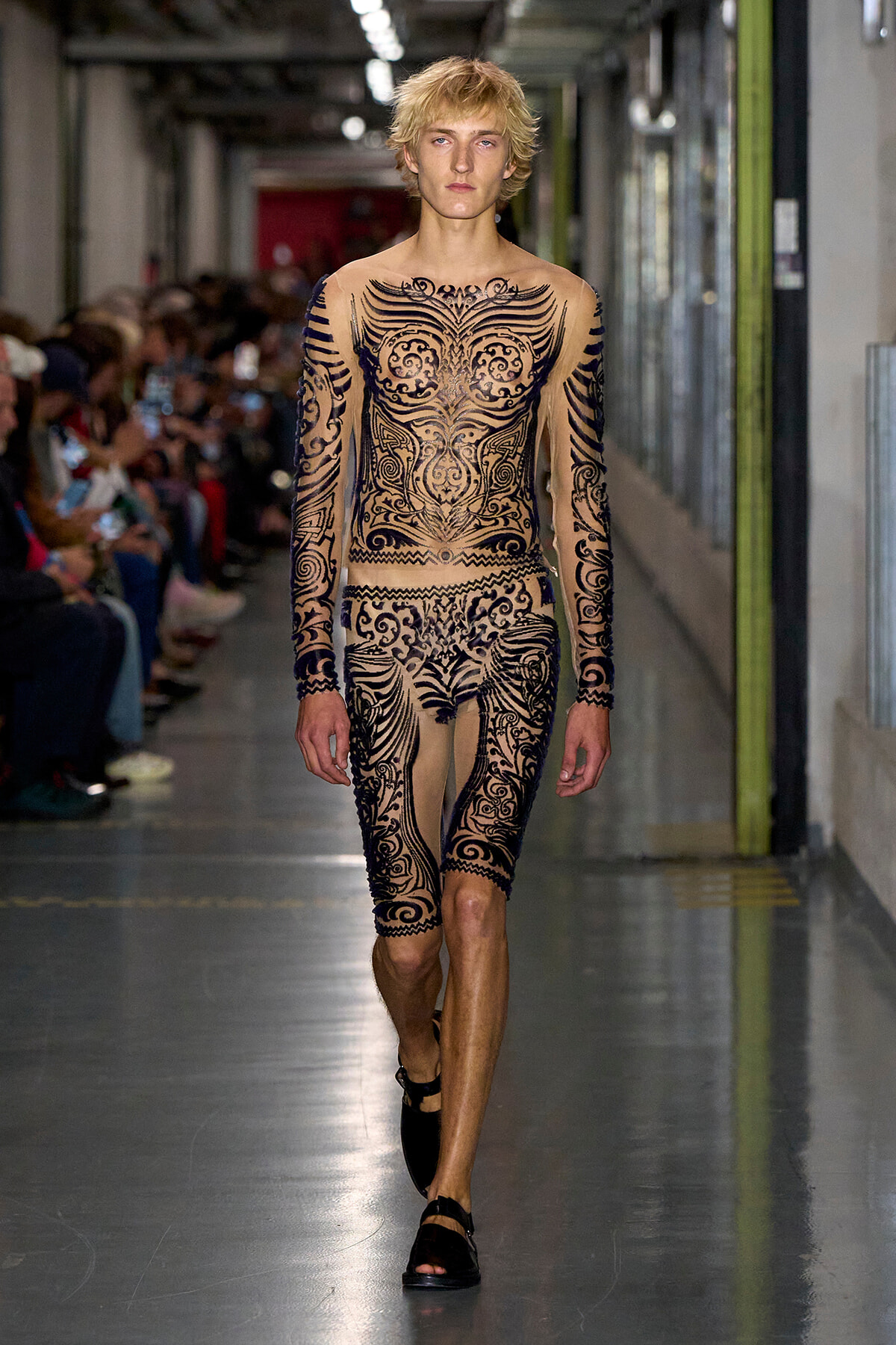 Male model walks the runway in a nude-toned sheer black lace bodysuit with ornate swirling embroidery.