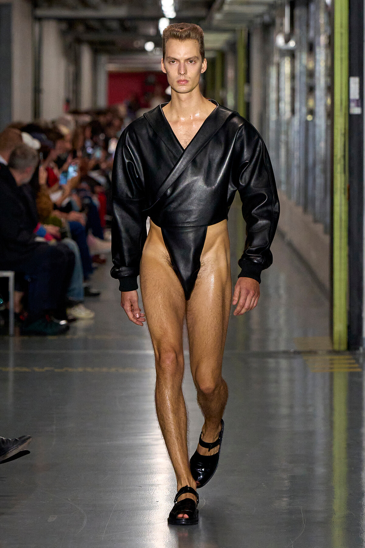 Male model walks a runway wearing a black leather-like wrap top with exaggerated shoulders and a high-cut black bottom, paired with black sandals.