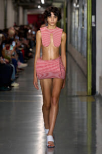 Runway model wearing a red-and-white striped crop top with a U-shaped cutout and a matching striped mini skirt, white sandals on a fashion show catwalk.