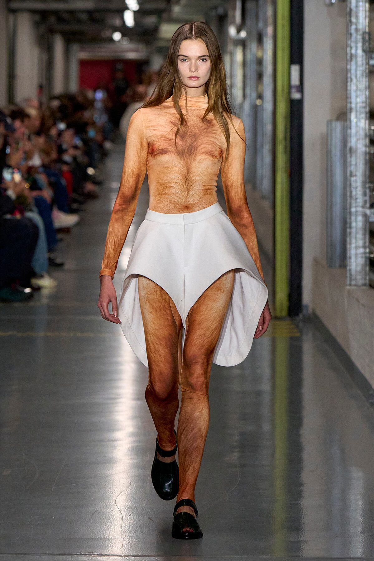 Female runway model walks a fashion show wearing a high-low white sculptural skirt, topless with visible body hair, black shoes; audience in background.