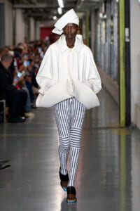Model on a runway wearing an oversized white coat with a large folded design, white sculptural hat, and black-and-white vertical striped leggings.