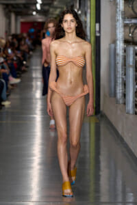 Model walks a runway in an orange-and-white striped bandeau bikini with knot details and matching side-tie bottoms, yellow sandals.