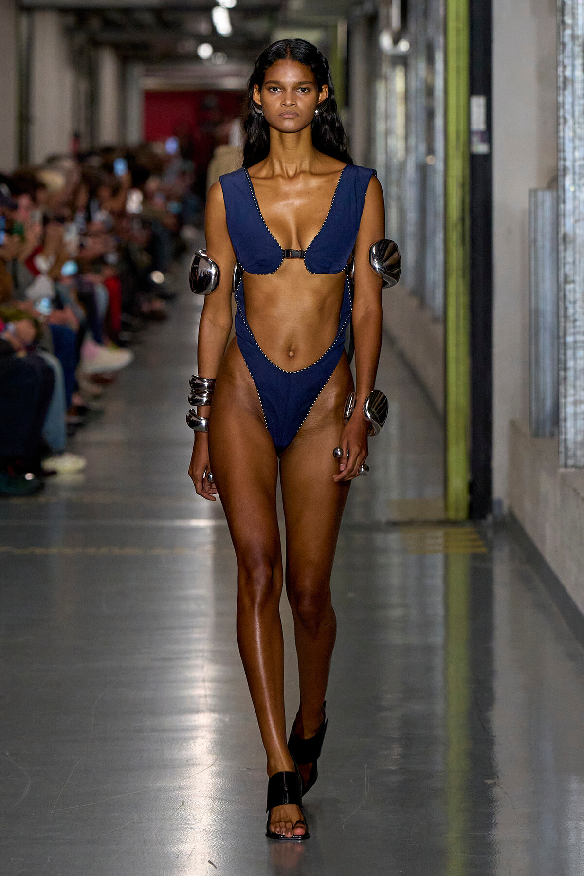 Model walks the runway in a navy blue bikini with metal armor on the upper arms and wrists, accessorized with bracelets during a fashion show in an industrial hallway.