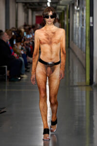 Model walks a runway wearing a full-body tan fur-patterned bodysuit, black belt, sunglasses, and sandals.