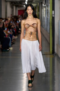 Model walks the runway with body paint on her chest and torso, wearing a white pleated midi skirt and green sandals, audience visible in the background.