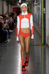 Runway model wearing a white ribbed hooded crop top with orange panels and matching high-cut orange bottoms, plus orange socks and sandals.
