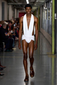 Male model walks a runway in a white draped one-piece outfit with a deep V, brown sandals, in an industrial-looking backstage hallway with seated audience on the sides.