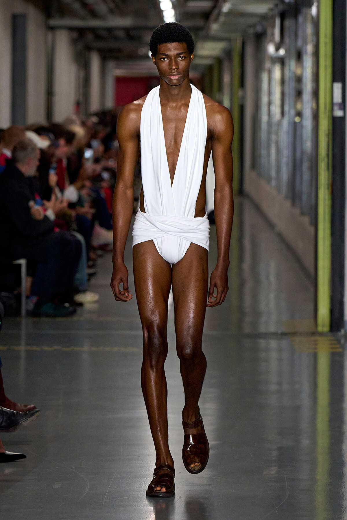 Male model walks a runway in a white draped one-piece outfit with a deep V, brown sandals, in an industrial-looking backstage hallway with seated audience on the sides.