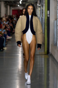 Model walks a runway wearing a beige oversized bomber jacket with a dark navy scarf over a white bodysuit and white ankle boots.