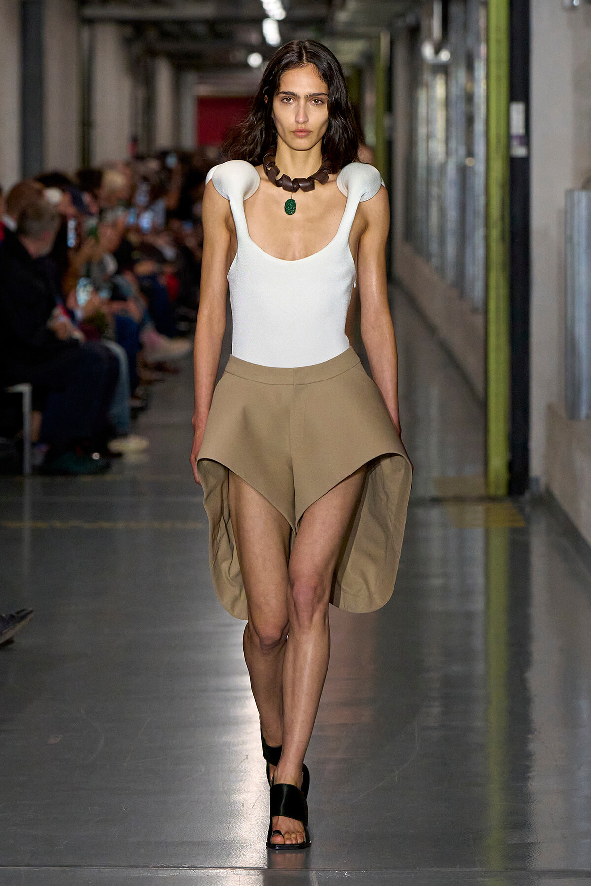 Model walking a runway in a white sculptural top and a taupe high-low skirt with a dramatic back hem, black sandals, and a chunky necklace.