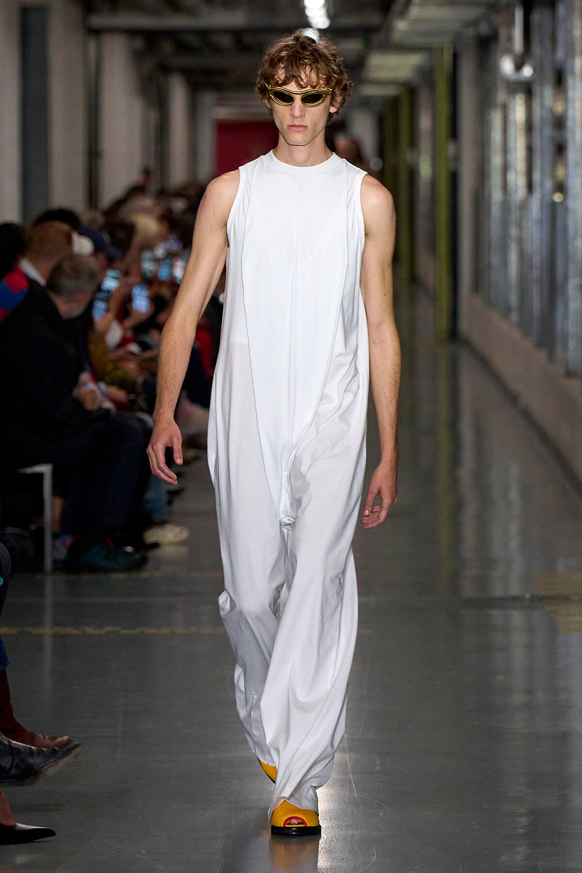 Male model walks a runway in a sleeveless white jumpsuit with yellow sandals and sunglasses.