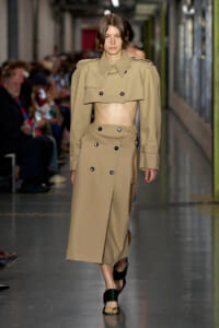 Female model walks a fashion runway in a beige cropped trench and matching high-waisted trench skirt with double-breasted buttons.