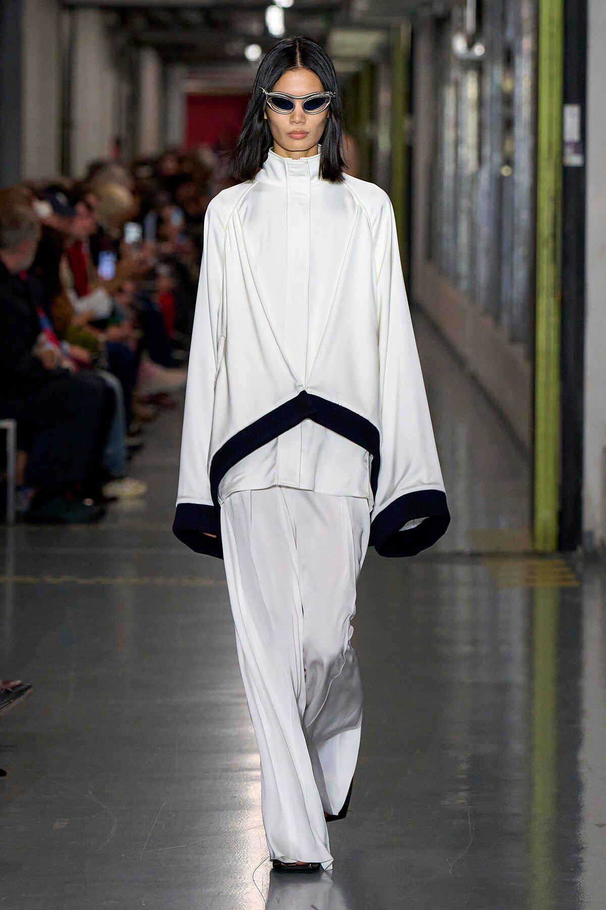Model walking a runway in an oversized white outfit with black trim at the cuffs and hem, wearing sleek sunglasses.