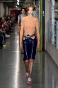 Model walks run way wearing orange-and-white striped long-sleeve top with hip cutouts, navy knee-length shorts, and white sandals with a wide belt.