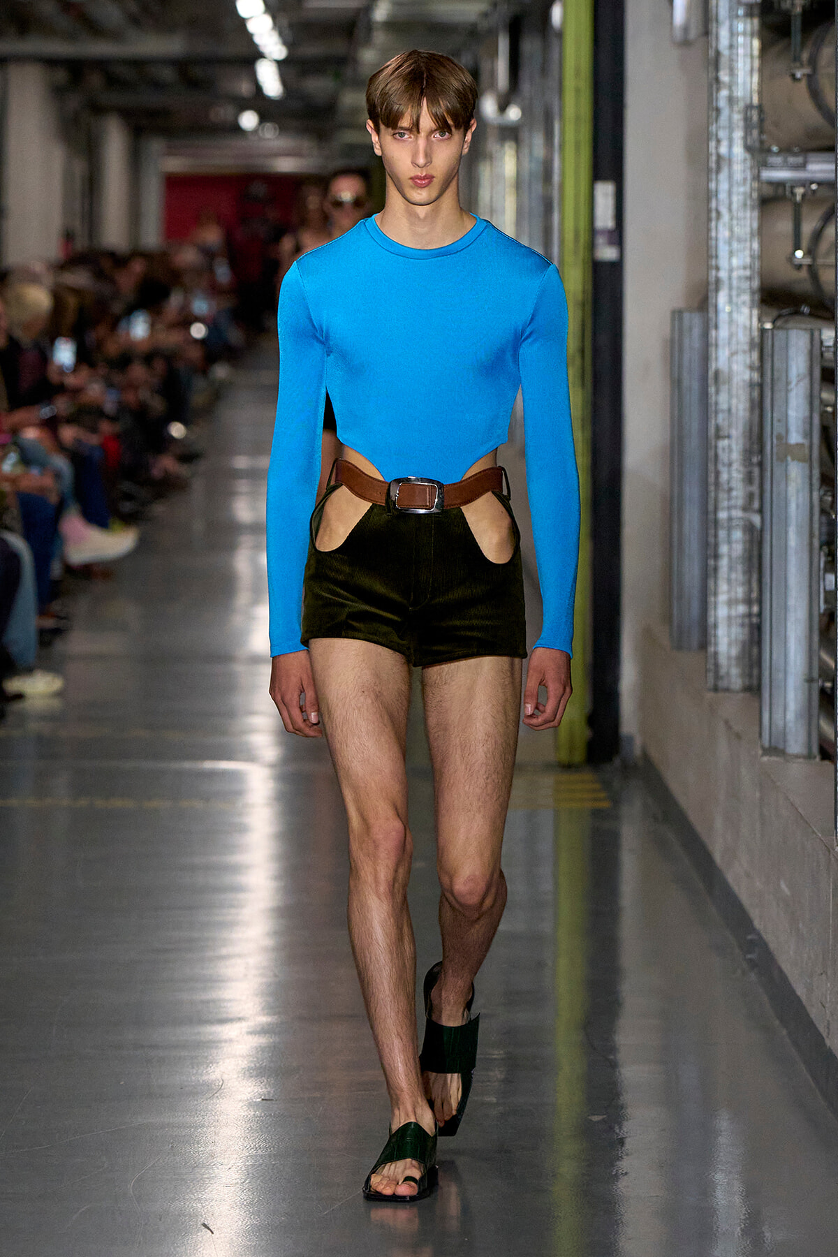 Male model on a runway wearing a bright blue long-sleeve top with waist cutouts, black shorts, a brown belt, and dark sandals.