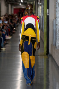 Model walking a runway in a bold color-blocked outfit with red, yellow, black, white top and blue/yellow pants, wearing oversized sunglasses.