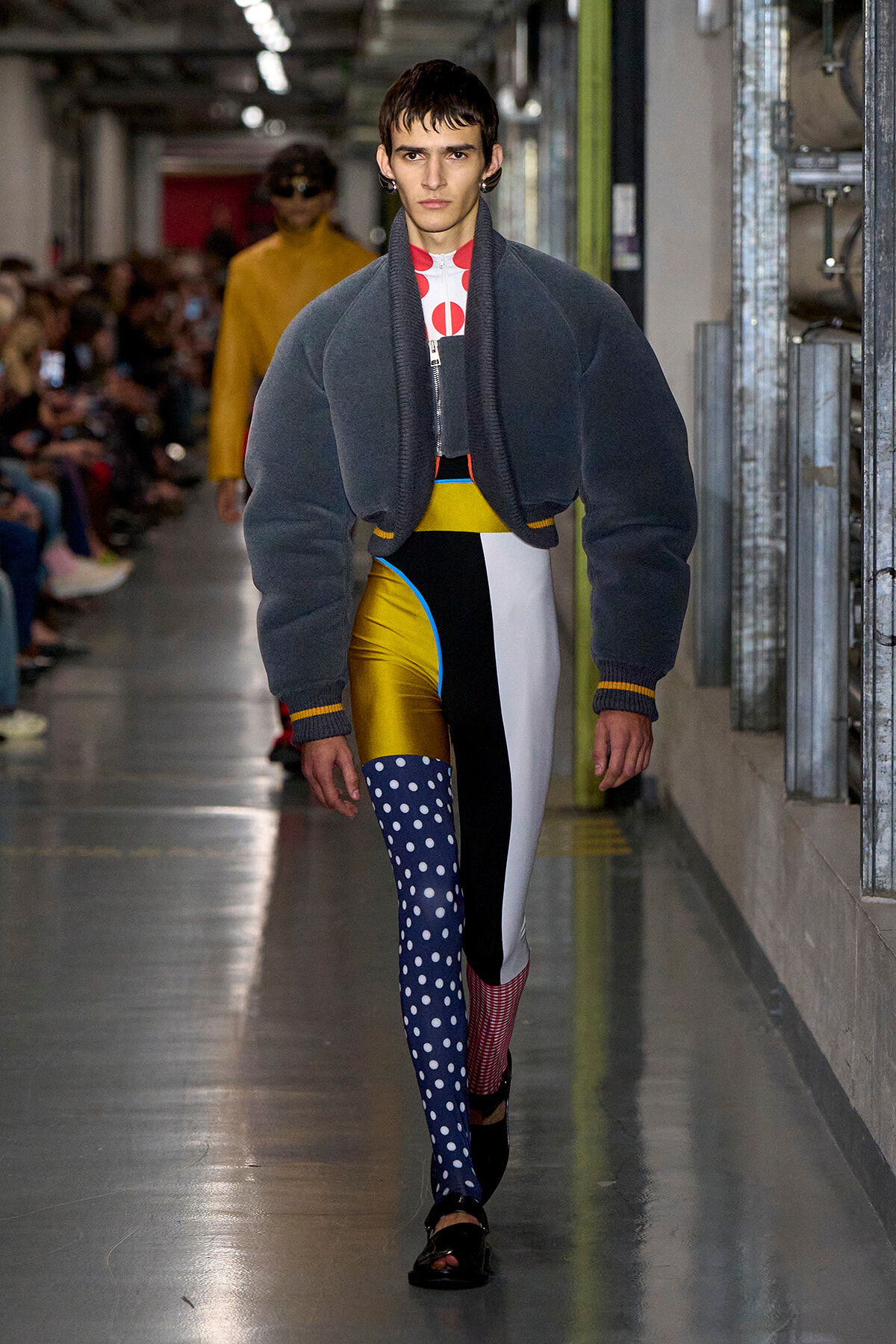 Model walks a runway in an oversized navy bomber-style jacket paired with bold color-blocked patchwork pants, featuring a blue polka-dot leg.