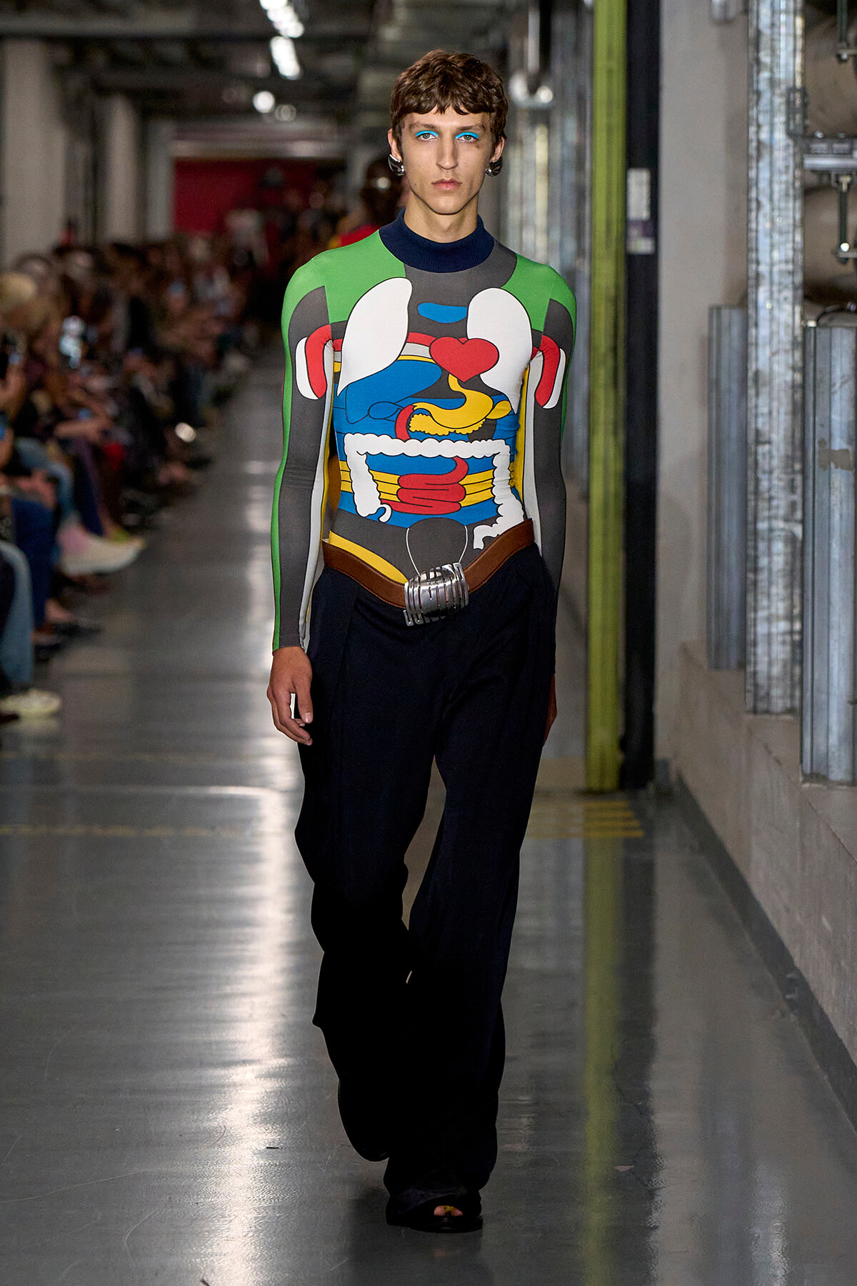 Model walking a runway in a bold, multicolored abstract bodysuit with green sleeves, paired with black wide-leg pants and a metallic belt in an industrial backstage setting.