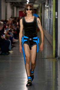 Model walking runway in a black sleeveless velvet top with a bright blue utilitarian harness and matching leg straps; wearing sunglasses.