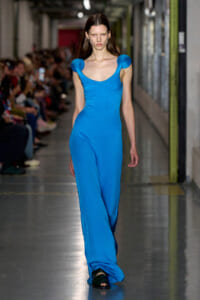 Female model walks a fashion show runway in a bright cobalt blue full-length jumpsuit with sculpted shoulder sleeves.]','Model wearing a vivid blue jumpsuit with rounded shoulder accents on a runway show.','Blue jumpsuit with cap-like shoulder details, model striding down the catwalk.','Full-length cobalt jumpsuit on a model during a fashion show.','Model in a striking blue jumpsuit with exaggerated shoulders on the runway.