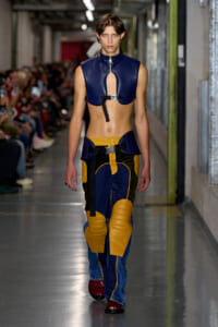 Model walking a fashion runway in a blue cropped leather top with a front zipper and color-blocked yellow/navy padded pants.