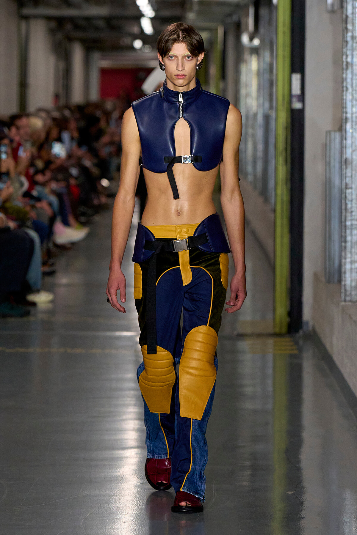 Model walking a fashion runway in a blue cropped leather top with a front zipper and color-blocked yellow/navy padded pants.