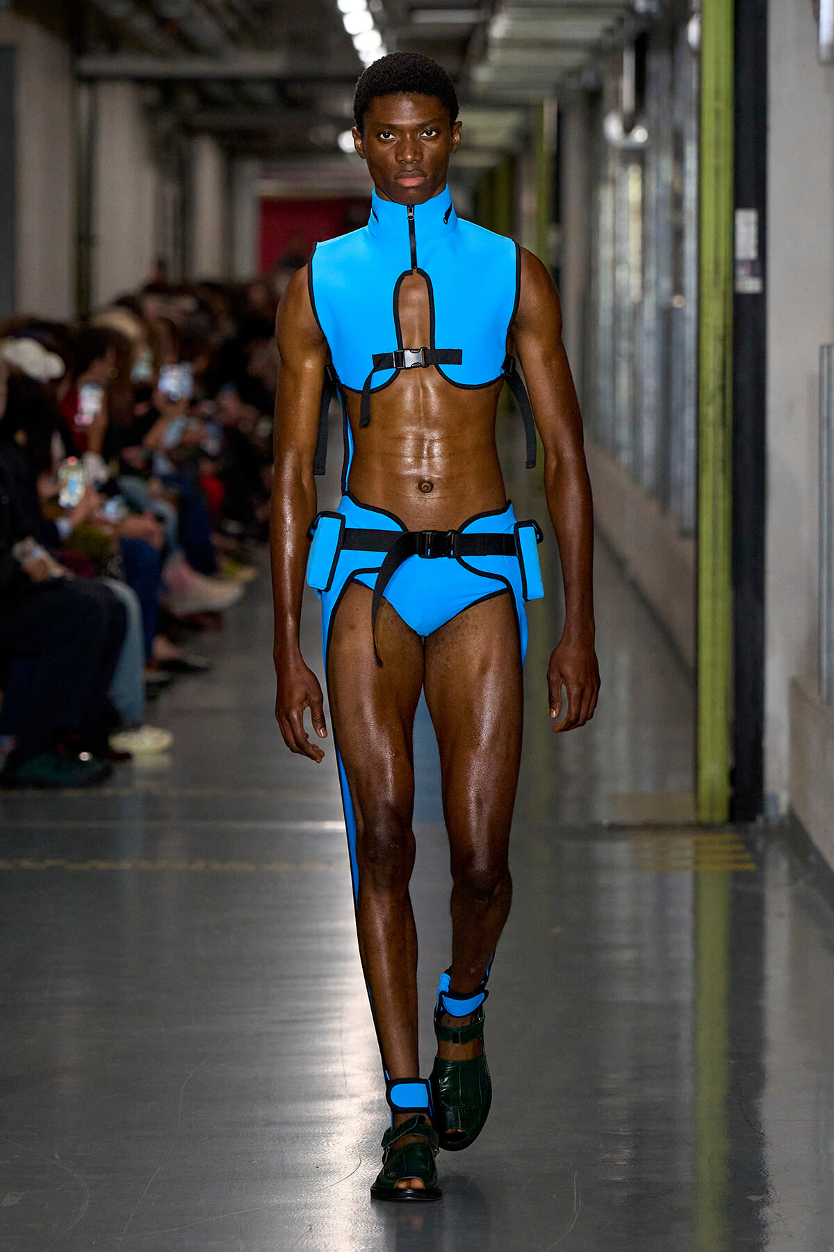 Male model on a fashion runway wearing a bright blue cutout harness with matching briefs and black straps.