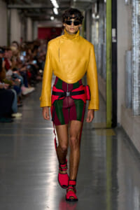 Model walking a runway in a mustard-yellow leather jacket with a high neck, paired with red and green color-block shorts and pink sandals, plus dark sunglasses.