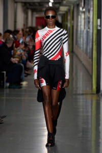 Runway model wearing a white, black, and red graphic striped top with black draped shorts and dark sunglasses, walking a fashion show hallway.
