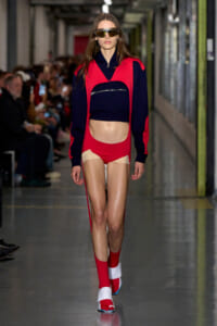 Model walking a runway in a red and navy color-block outfit: cropped navy top with a zip pocket under a red overlay and matching red shorts, red socks, and white sandals, wearing mirrored sunglasses.