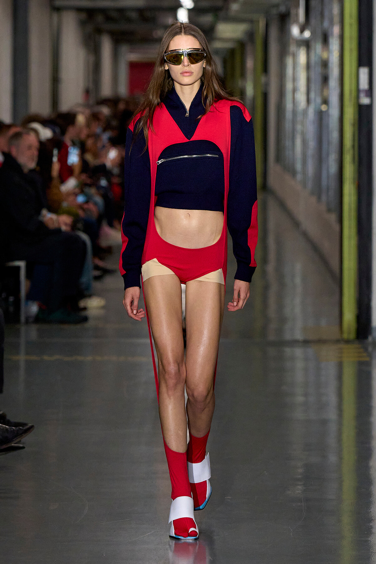 Model walking a runway in a red and navy color-block outfit: cropped navy top with a zip pocket under a red overlay and matching red shorts, red socks, and white sandals, wearing mirrored sunglasses.
