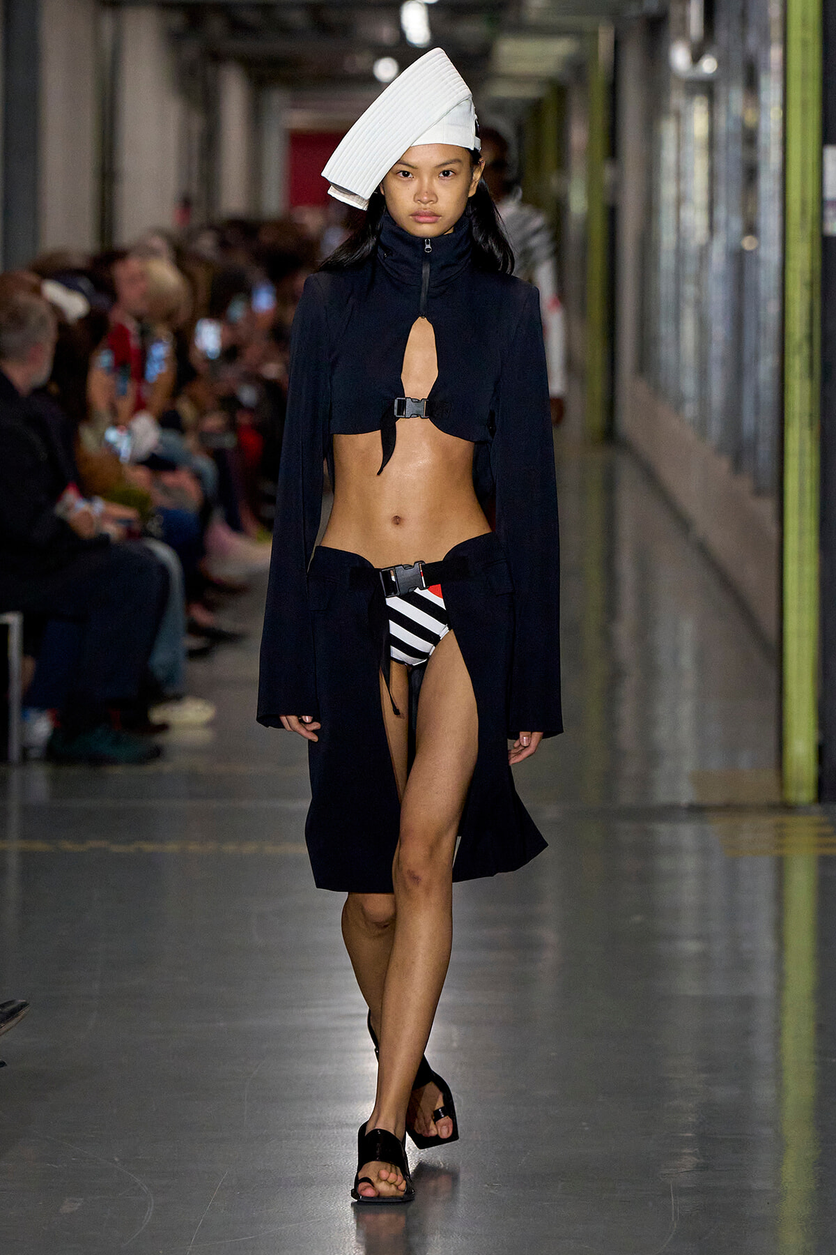 Model on a runway wearing a black cropped coat with a center keyhole, striped black-and-white bikini bottoms, and a large white sculptural hat.