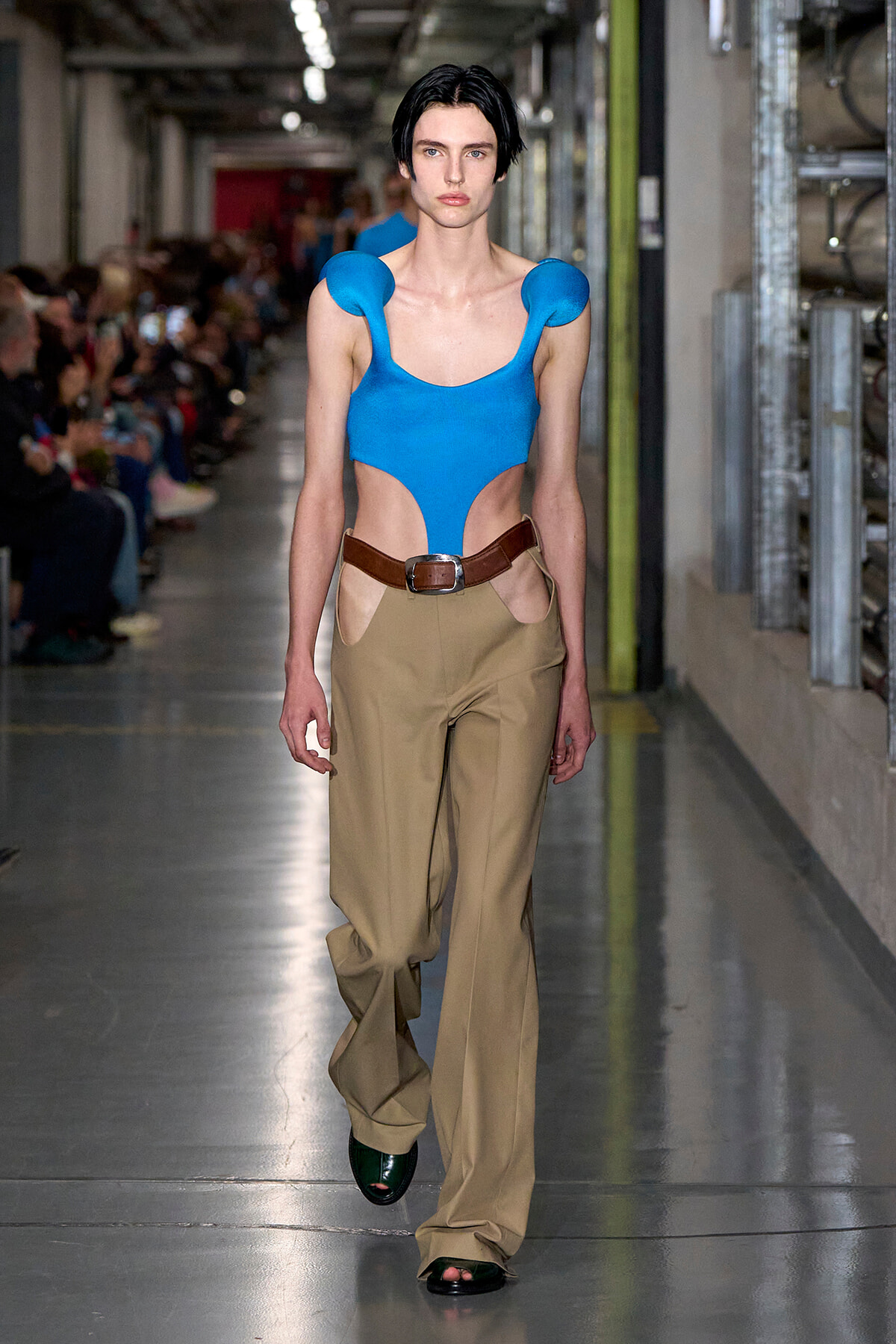 Model walking a runway in a bright blue cutout top with a brown belt and tan wide-leg pants.”],