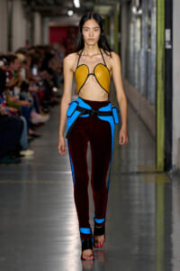 Model walking runway in a mustard-yellow open-front halter top and burgundy velvet pants with blue harness detailing, black sandals.