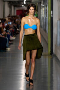 Model walks a runway in a bright blue strapless bandeau top and an olive green high-low skirt, black sandals.