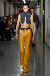 Model walking a runway in a sleeveless gray high-neck top with front cutouts paired with mustard leather pants and a burgundy belt.