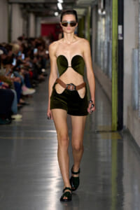 Model walking a fashion show in an olive-green strapless top with heart-shaped cups and matching shorts, brown belt, and sunglasses.