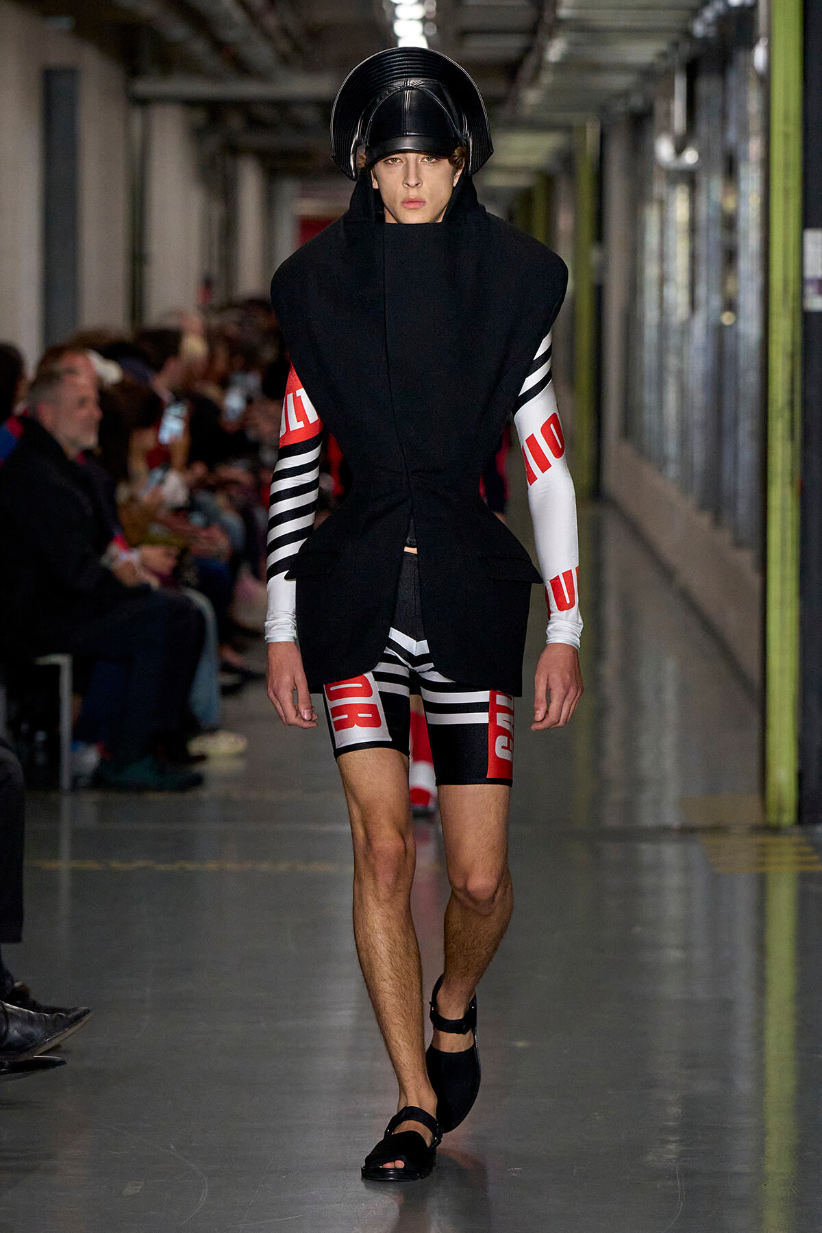Runway model in a futuristic black sculpted coat with striped sleeves and bold red text, wearing a black helmet headpiece.