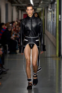 Male model walking a runway in a black leather shirt and avant-garde black-and-white skirt with white piping and high-cut sides.
