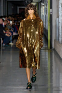 Model walks down a runway wearing a long, oversized gold sequin coat with hands in pockets and dark green sandals.
