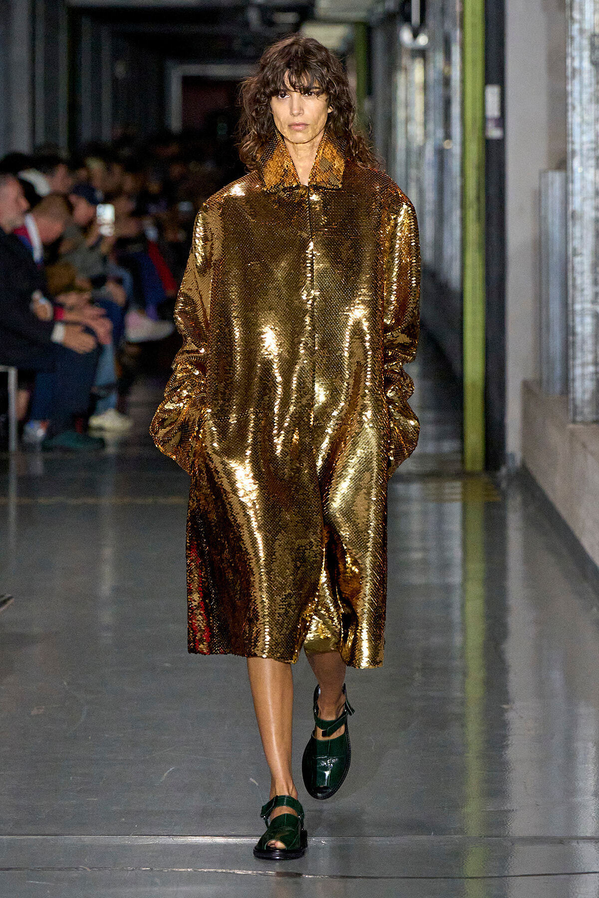 Model walks down a runway wearing a long, oversized gold sequin coat with hands in pockets and dark green sandals.