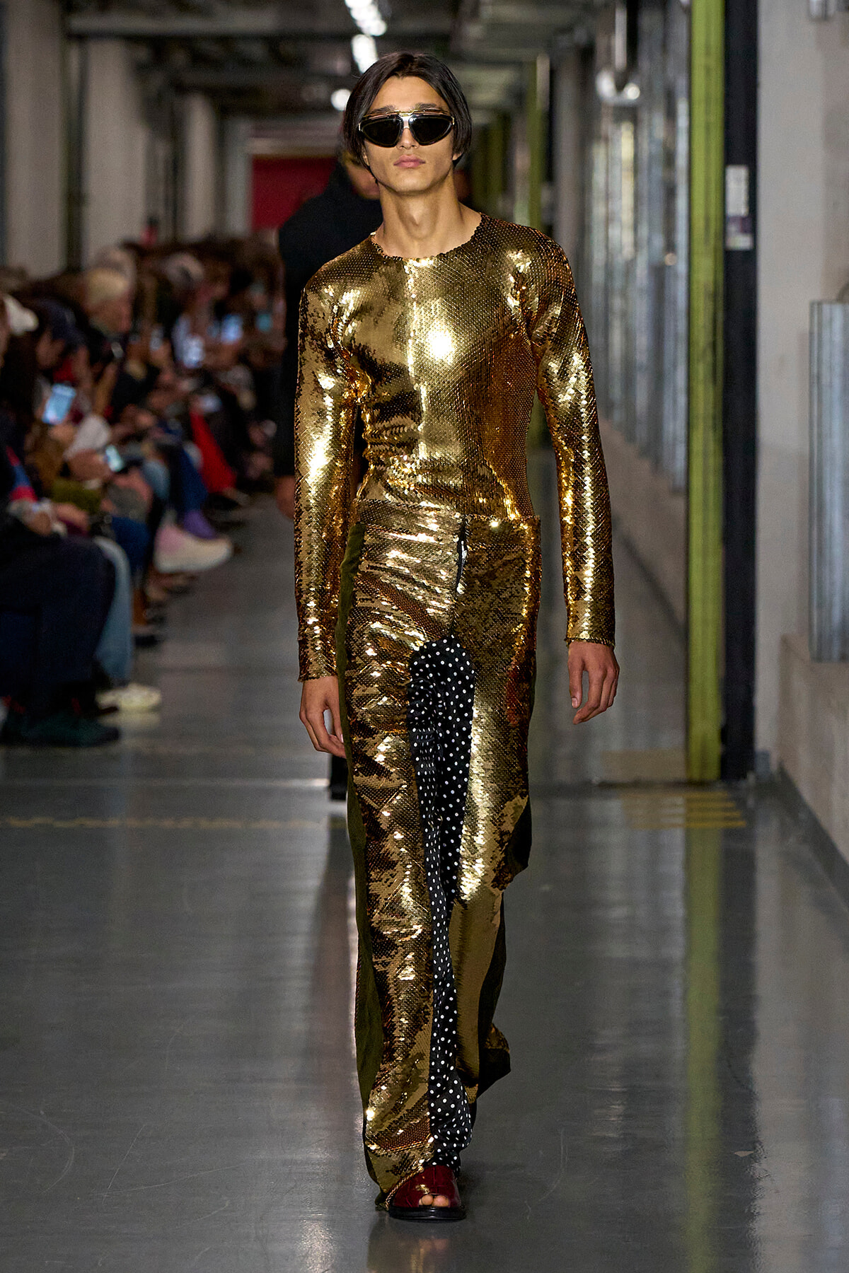 Model walking a fashion show in a full gold sequin outfit with sunglasses, on a runway lined with spectators.